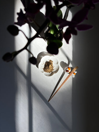 A minimalist tabletop scene with a small white dish holding brown powder, a pair of wooden chopsticks resting beside it, and dark purple orchid flowers hanging over the frame against a brightly lit white wall with strong shadows.